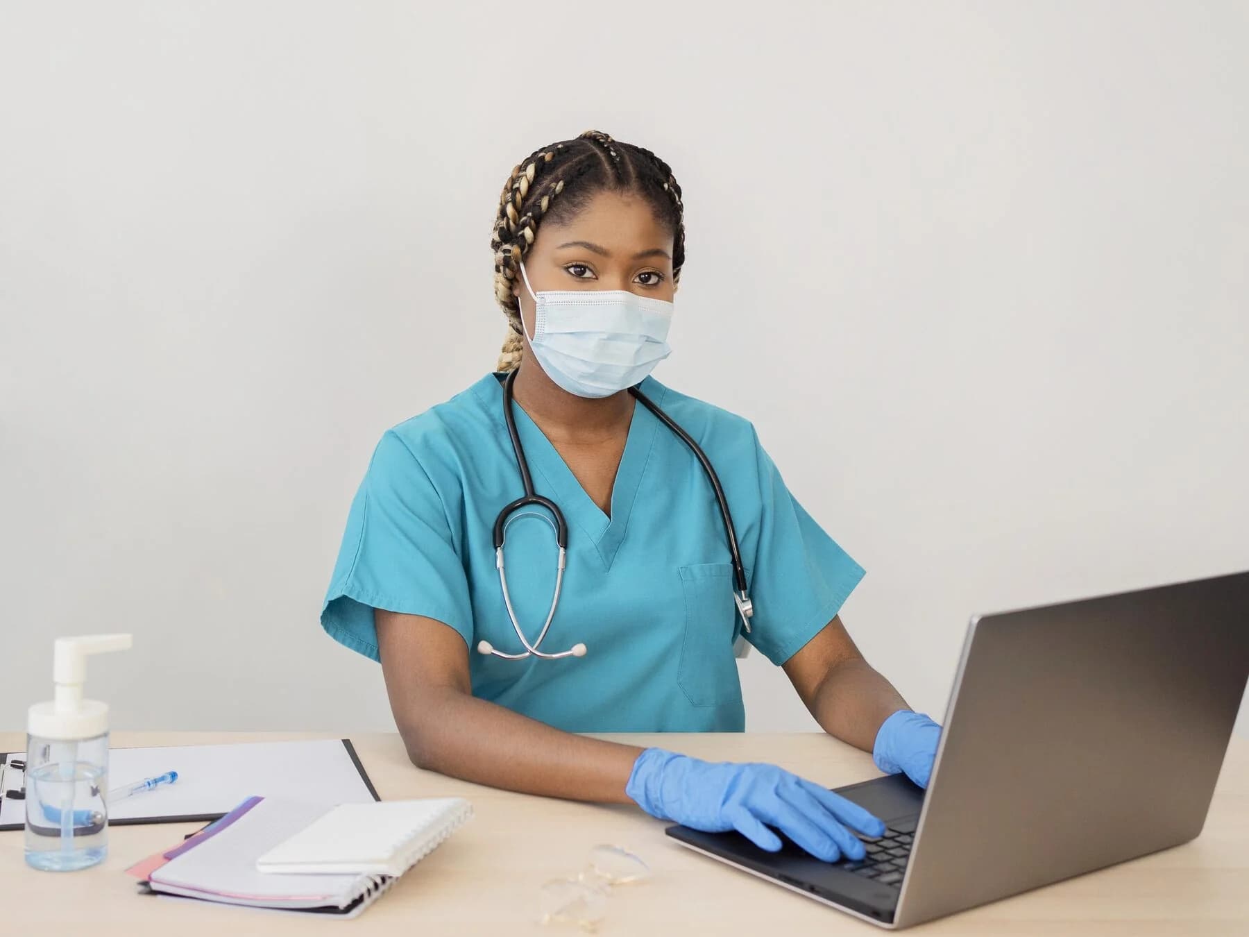 Healthcare worker in scrubs and a face mask working on a laptop at a clinic desk