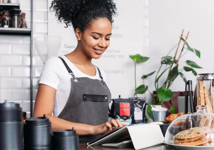 Smiling café worker in an apron entering an order on a tablet at the counter
