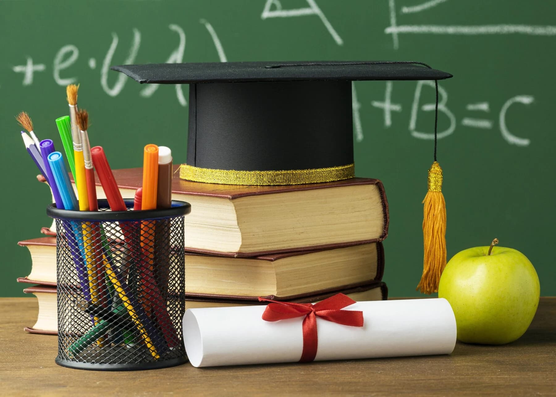 Graduation cap on a stack of books with pencils, a diploma and an apple in front of a chalkboard